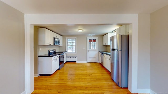 kitchen featuring dark countertops, light wood finished floors, radiator heating unit, appliances with stainless steel finishes, and white cabinetry