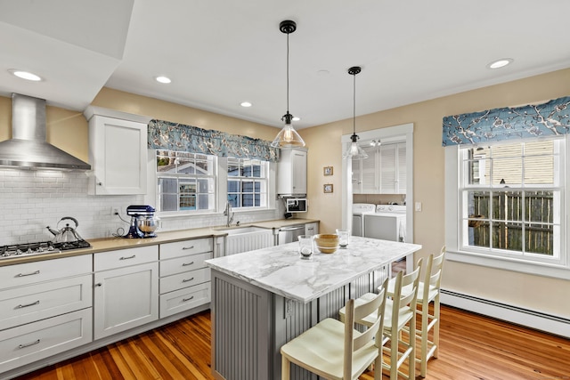 kitchen featuring wood finished floors, washing machine and dryer, appliances with stainless steel finishes, wall chimney range hood, and baseboard heating