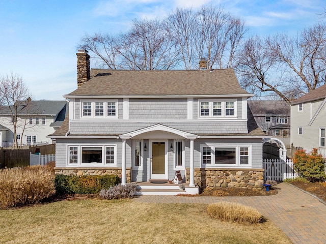 shingle-style home with stone siding, a chimney, a front lawn, and fence