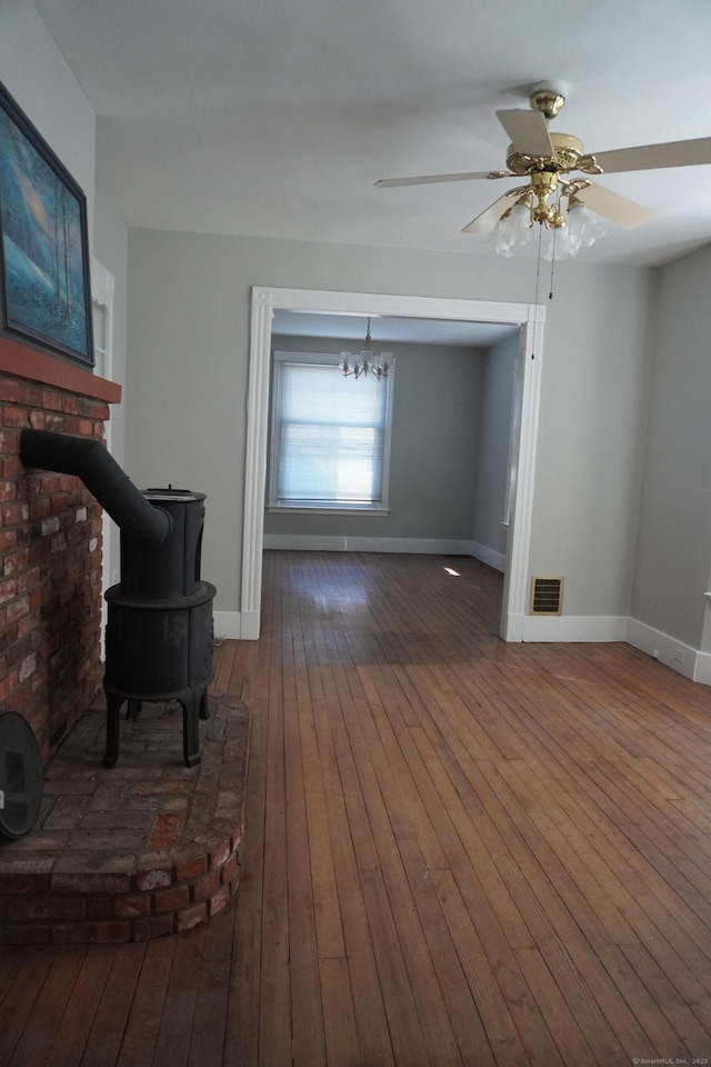 unfurnished living room with hardwood / wood-style floors, visible vents, baseboards, a wood stove, and ceiling fan with notable chandelier