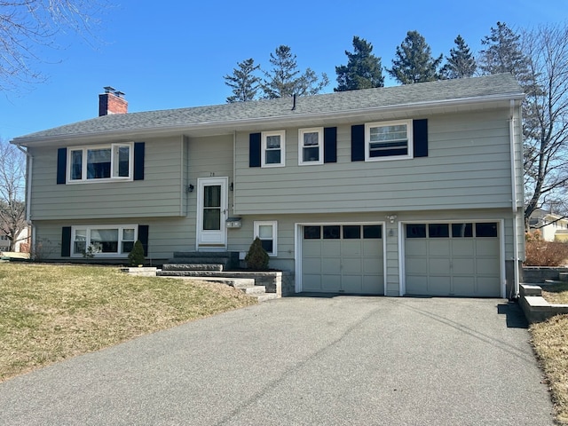 raised ranch with driveway, a shingled roof, a front yard, an attached garage, and a chimney