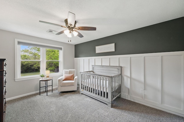 carpeted bedroom with a nursery area, a textured ceiling, ceiling fan, and wainscoting