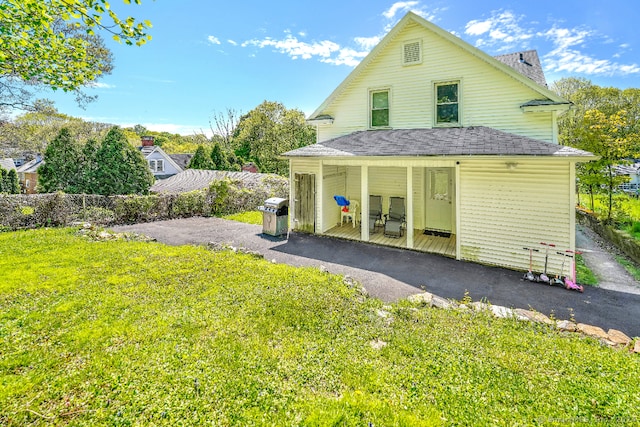 rear view of property with fence, driveway, roof with shingles, a porch, and a yard