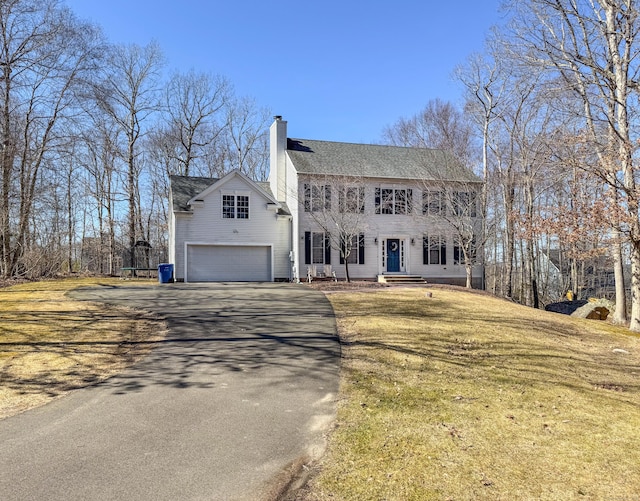 colonial-style house featuring a front yard, driveway, an attached garage, a shingled roof, and a chimney
