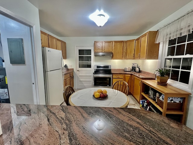 kitchen featuring electric panel, stainless steel electric range, under cabinet range hood, brown cabinetry, and freestanding refrigerator