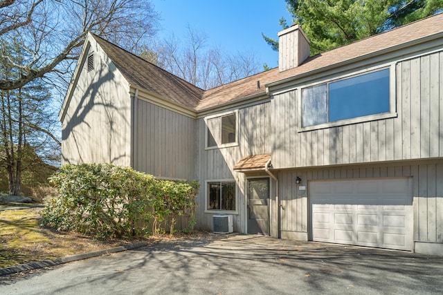 view of front of home with driveway, central AC, an attached garage, a shingled roof, and a chimney