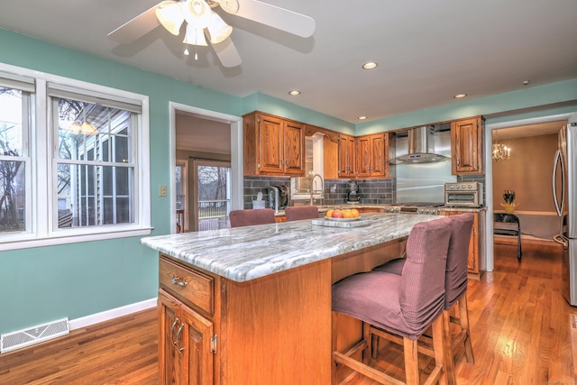 kitchen with light wood finished floors, wall chimney exhaust hood, brown cabinets, visible vents, and decorative backsplash