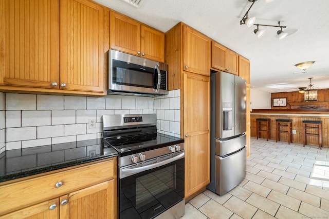kitchen with backsplash, light tile patterned floors, dark stone countertops, appliances with stainless steel finishes, and brown cabinetry
