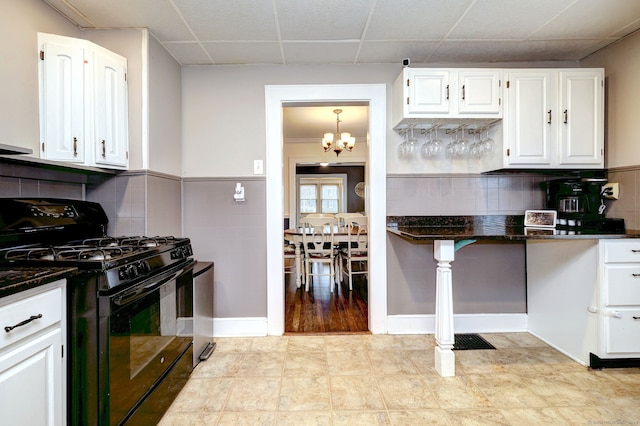 kitchen with dark stone countertops, a drop ceiling, black gas range oven, white cabinets, and a notable chandelier