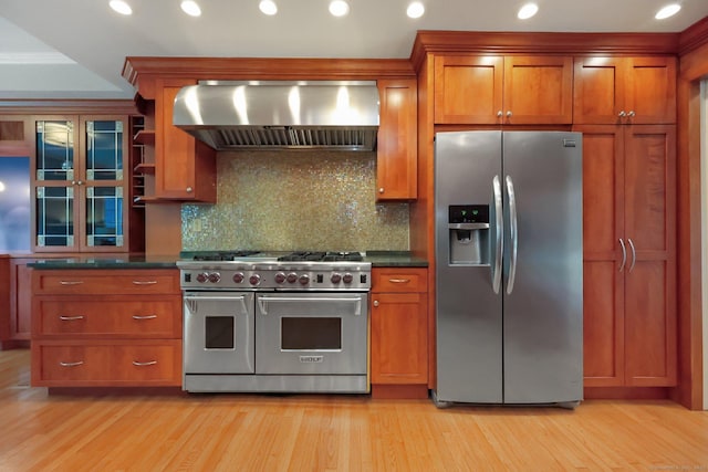 kitchen featuring appliances with stainless steel finishes, wall chimney range hood, brown cabinetry, dark countertops, and recessed lighting