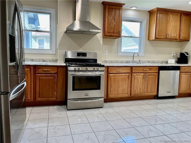 kitchen featuring light stone counters, a sink, wall chimney exhaust hood, brown cabinets, and stainless steel appliances