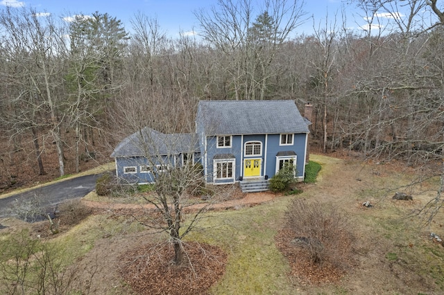 view of front of home with a shingled roof, a chimney, driveway, and a wooded view
