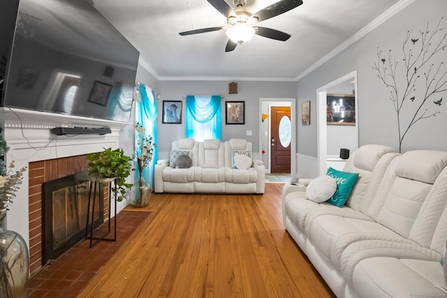 living room featuring wood finished floors, a ceiling fan, wainscoting, a brick fireplace, and crown molding