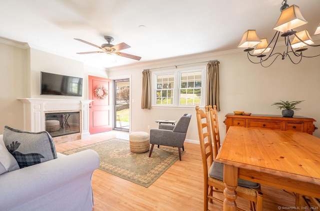 living area with crown molding, ceiling fan with notable chandelier, a glass covered fireplace, light wood finished floors, and baseboards