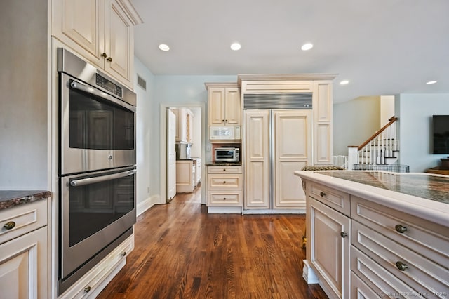 kitchen with dark stone countertops, double oven, paneled fridge, cream cabinetry, and dark wood-style floors