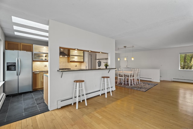 kitchen featuring stainless steel fridge with ice dispenser, wall chimney range hood, a baseboard radiator, and a breakfast bar area