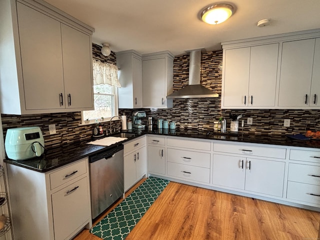 kitchen featuring wall chimney range hood, light wood-style floors, decorative backsplash, stainless steel dishwasher, and a sink