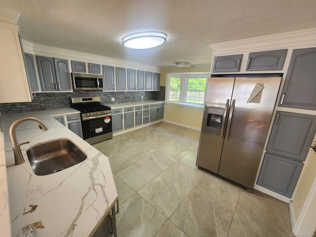 kitchen with light stone counters, a sink, gray cabinets, stainless steel appliances, and backsplash