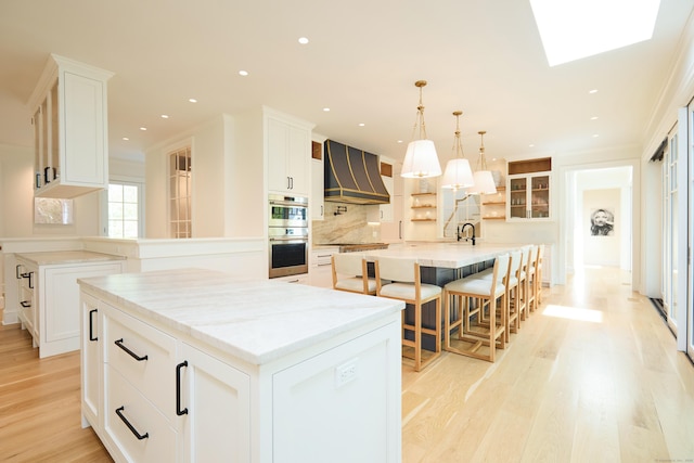 kitchen featuring double oven, a center island with sink, light wood-type flooring, tasteful backsplash, and custom exhaust hood