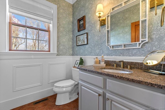 bathroom featuring visible vents, toilet, vanity, wainscoting, and wallpapered walls