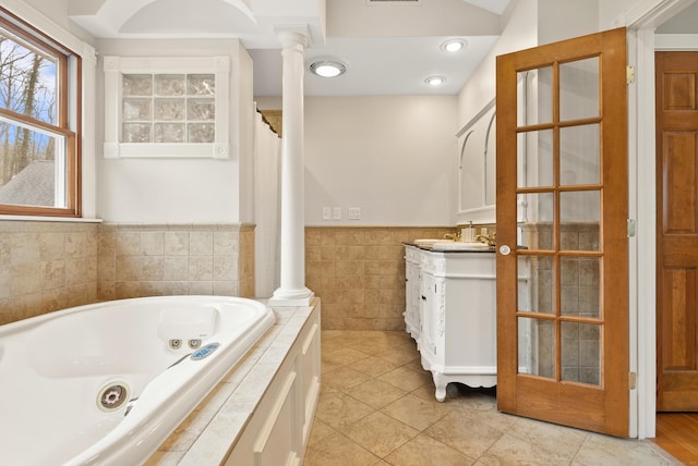 full bath featuring vanity, wainscoting, tile walls, a jetted tub, and ornate columns