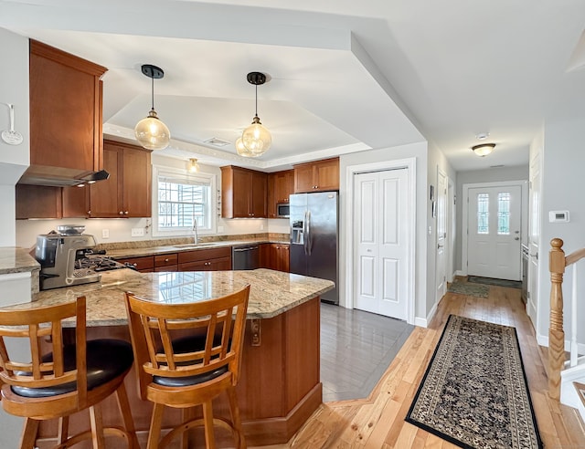 kitchen featuring stainless steel appliances, a peninsula, a sink, brown cabinetry, and a raised ceiling