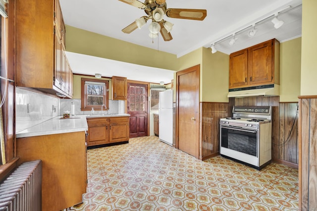 kitchen featuring white appliances, under cabinet range hood, brown cabinets, wainscoting, and radiator heating unit