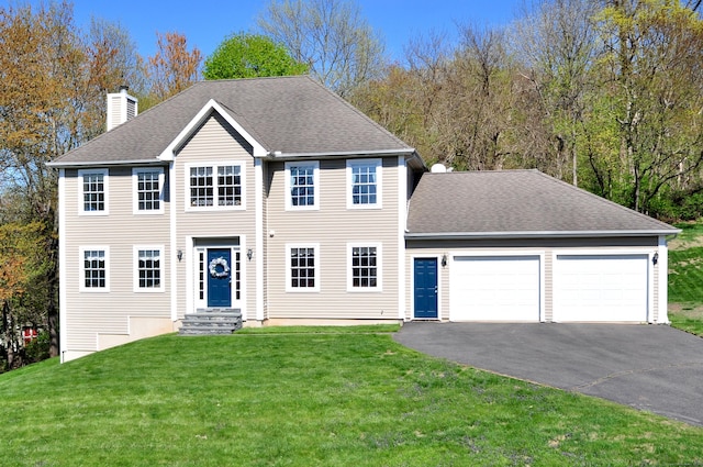 colonial-style house featuring roof with shingles, a front lawn, aphalt driveway, a garage, and a chimney