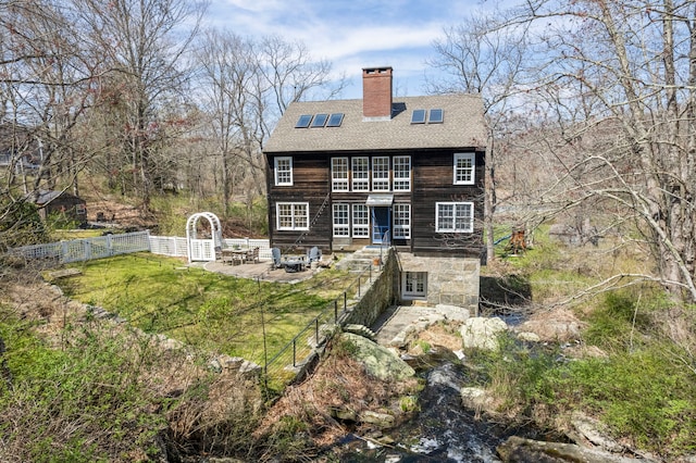 rear view of house with a chimney, a shingled roof, an outdoor fire pit, a lawn, and fence