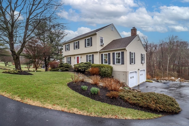 colonial-style house featuring a garage, a front lawn, a chimney, a shingled roof, and driveway