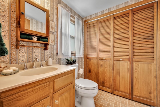 bathroom featuring a closet, toilet, brick floor, vanity, and wallpapered walls