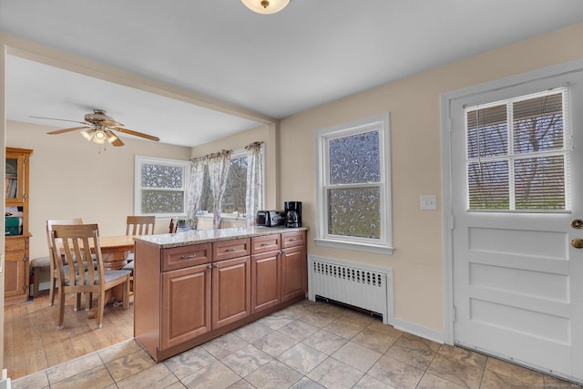 kitchen featuring ceiling fan, a peninsula, brown cabinets, radiator, and light stone countertops