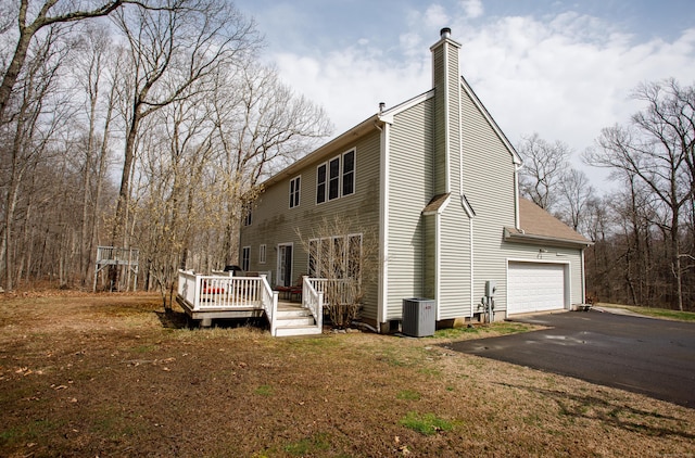back of house featuring driveway, a chimney, a deck, an attached garage, and central AC