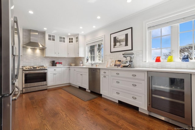 kitchen with beverage cooler, wall chimney exhaust hood, stainless steel appliances, white cabinetry, and backsplash
