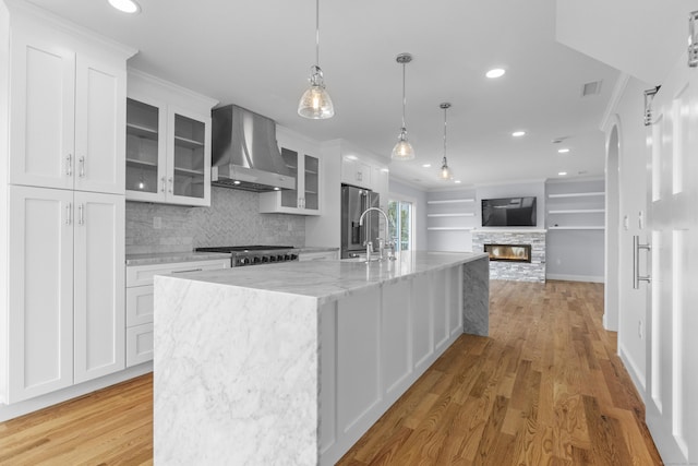 kitchen featuring white cabinets, a sink, wall chimney range hood, an island with sink, and stove
