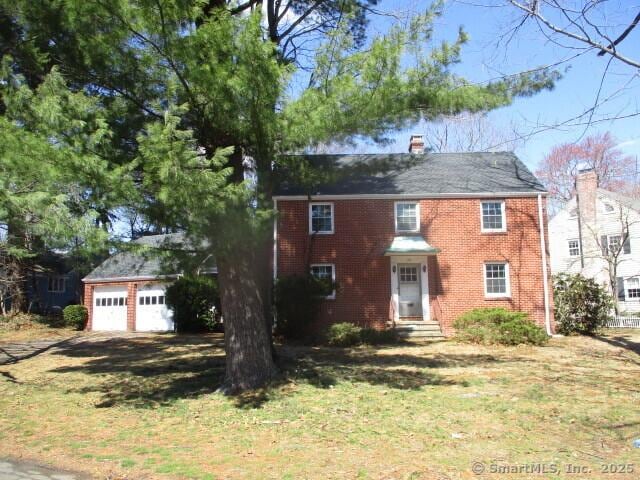colonial-style house featuring brick siding, a front lawn, a chimney, and a detached garage
