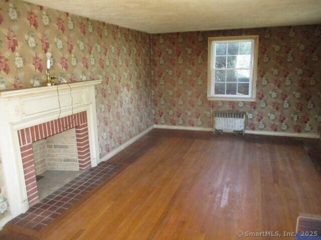 unfurnished living room with radiator, wood finished floors, a brick fireplace, wallpapered walls, and baseboards