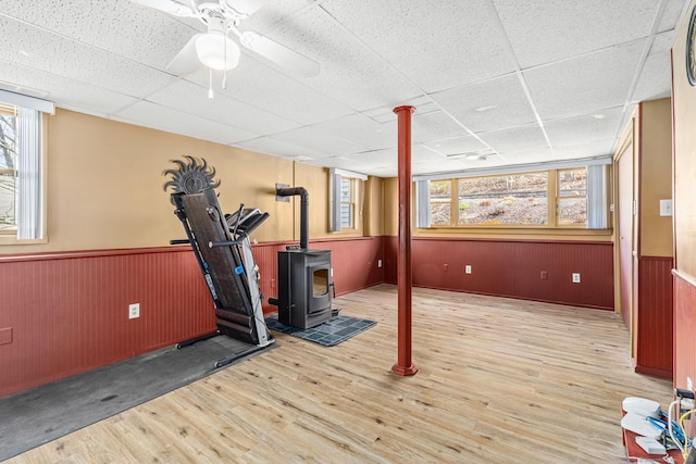 workout room with a wood stove, ceiling fan, a paneled ceiling, wood finished floors, and a wainscoted wall