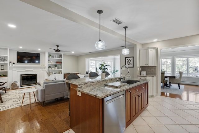 kitchen with a sink, a glass covered fireplace, stainless steel dishwasher, visible vents, and a center island with sink