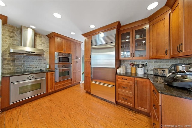 kitchen featuring light wood-style floors, wall chimney range hood, brown cabinets, tasteful backsplash, and appliances with stainless steel finishes