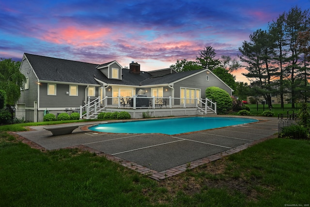 back of house at dusk featuring a wooden deck, an outdoor pool, roof with shingles, a yard, and a chimney