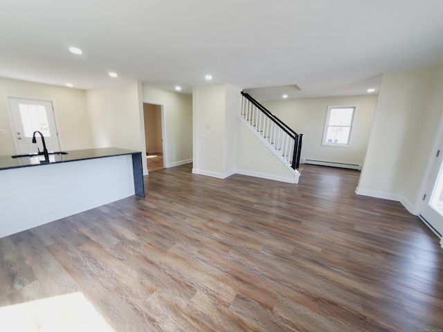 unfurnished living room featuring recessed lighting, wood finished floors, a baseboard radiator, a sink, and stairway