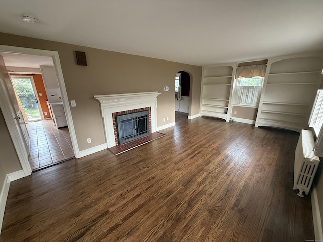 unfurnished living room with arched walkways, baseboards, built in shelves, and dark wood-type flooring