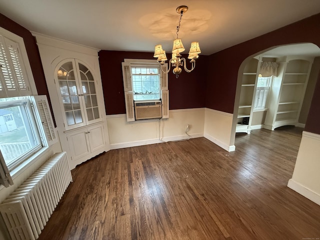 unfurnished dining area featuring radiator, cooling unit, arched walkways, an inviting chandelier, and dark wood finished floors