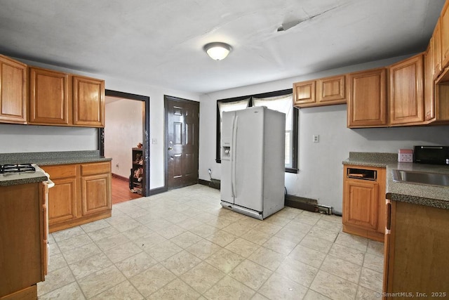 kitchen with a sink, dark countertops, baseboards, white refrigerator with ice dispenser, and brown cabinets