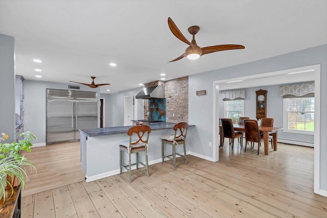 kitchen featuring wall chimney exhaust hood, a ceiling fan, dark countertops, a baseboard radiator, and built in fridge