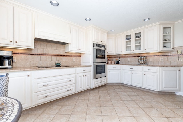 kitchen with light stone counters, tasteful backsplash, glass insert cabinets, stainless steel double oven, and black electric cooktop