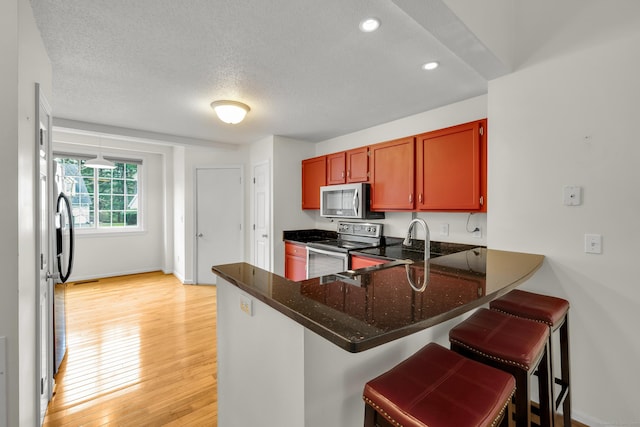 kitchen with a textured ceiling, appliances with stainless steel finishes, a peninsula, dark stone counters, and light wood-style flooring