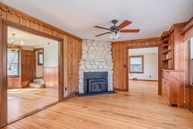 unfurnished living room featuring light wood finished floors, wooden walls, crown molding, baseboards, and ceiling fan with notable chandelier