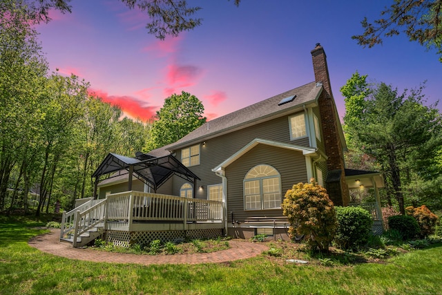 back of property at dusk with a deck, a gazebo, a chimney, and a lawn